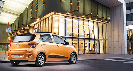 Side-rear view of orange Grandi10 passing by the glass building